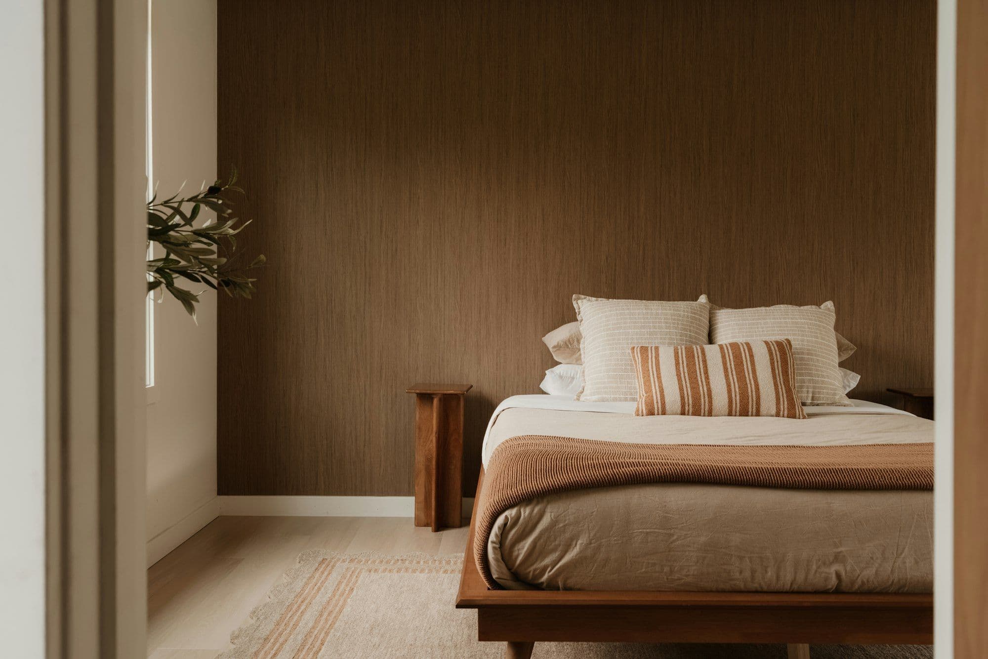 Slaapkamer in warme bruin- en zandtinten met een gestreepte wandbekleding, beige beddengoed, een plaid aan het voeteneinde en een vloerkleed.